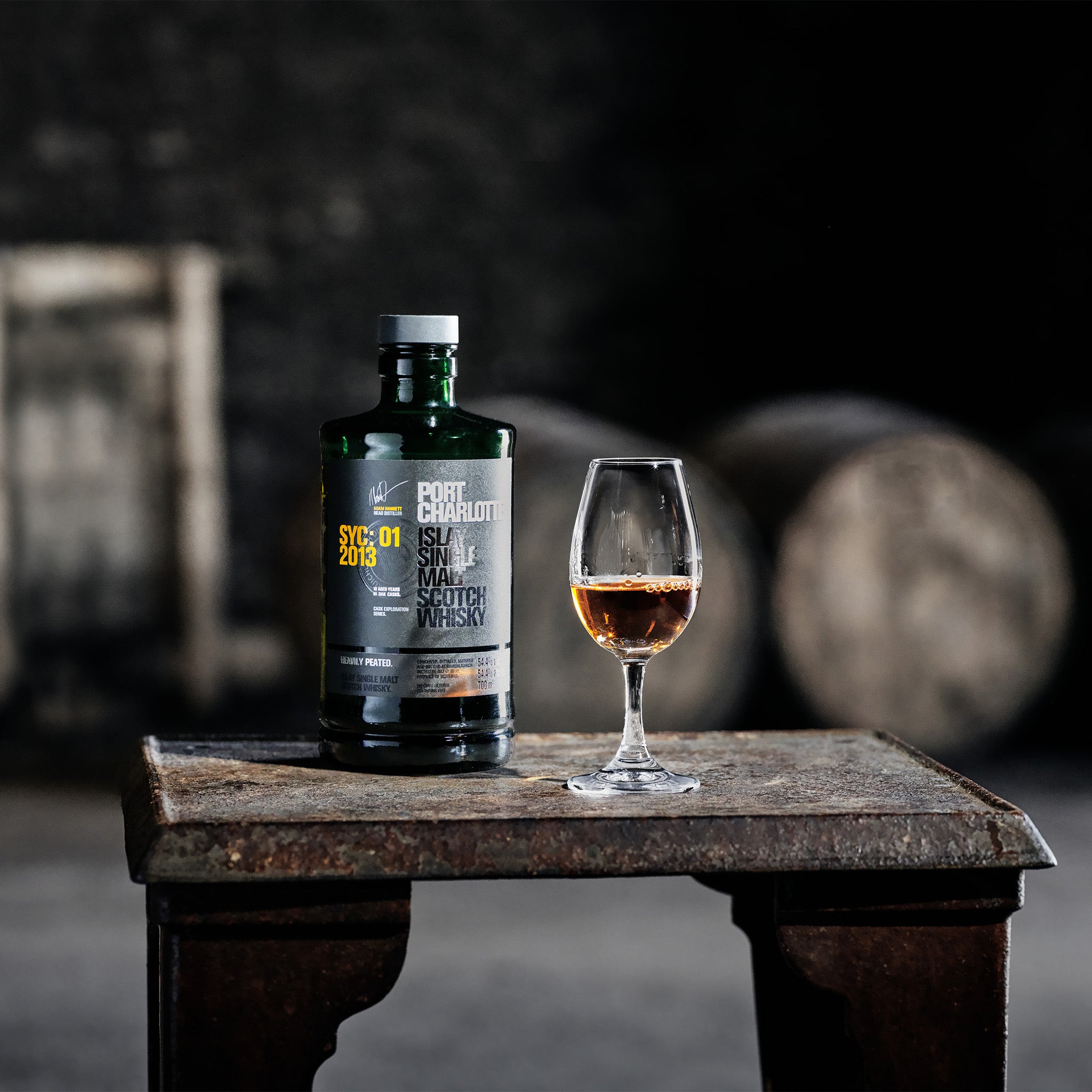 A bottle of Port Charlotte SYC:01 2013 whisky stands beside a glass filled with whisky, set against a background of casks in a warehouse.